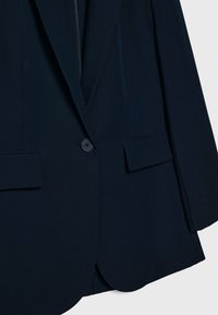 Mörk marinblå blazer med en knappförslutning, två framfickor med flik och slagkrage, uppvisad mot en enkel bakgrund.