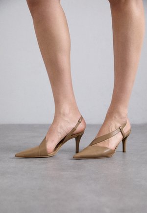 Nogi w beżowych szpilkach typu slingback z wąskim noskiem i średniej wysokości obcasach typu szpilka stojące na szarej powierzchni.