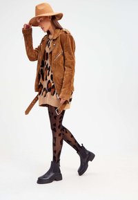 Tan suede moottoripyöräjacketti leopardikuvioisen mekon ja mustien kuosisukkahousujen päällä, yhdistettynä mustiin nilkkabootseihin ja vaaleanruskeaan huopahatun.