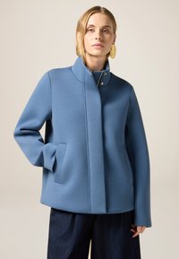 Giacca blu, con texture, dal colletto alto, tasche frontali e design minimalista. Presenta bottoni a scatto dorati e una vestibilità rilassata.