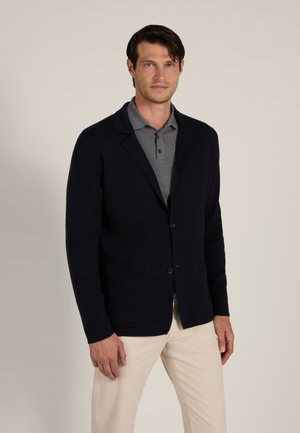 Man die een donkere blazer draagt over een grijs overhemd met kraag en lichtbeige broek, staand tegen een effen lichte achtergrond.