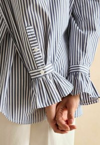 Triibuline pluus sinise ja valge vertikaalse triibuga, millel on volangvarrukad, nööbi sulgemised ja mugav lõige. Puuvillane materjal, sile tekstuur.