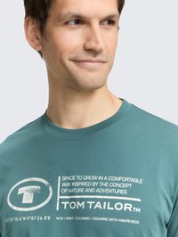 Teal katoen t-shirt met een ronde hals, met witte grafische tekst en logo op de voorkant, minimalistisch ontwerp.
