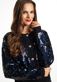 Juodas megztinis, puošiamas mėlynais ir sidabriniais blizgučiais, su apvaliais metaliniais mygtukais ir gumbuotomis rankovėmis, suteikiančiomis tekstūrinį akcentą.