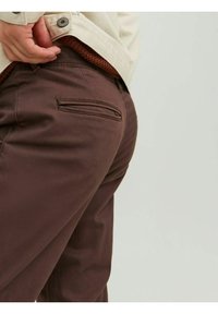 Pantaloni di cotone marroni con una vestibilità slim, dotati di una tasca posteriore e una sottile texture. La vita è abbinata a una camicia di colore chiaro.
