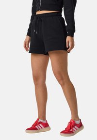 Person trägt schwarze Shorts mit Kordelzug, cropped schwarze Jacke, weiße Socken und rote Adidas-Sneaker vor einem schlichten Hintergrund.