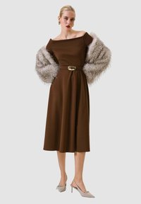 Vestito marrone con scollo alla schiena e vita stretta, accompagnato da uno scialle grigio soffice. Tacchi beige a punta con dettaglio a fiocco.