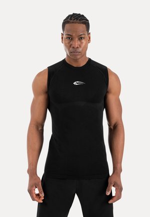 Uomo muscoloso con capelli intrecciati indossa una maglietta atletica nera senza maniche con logo bianco, in piedi davanti a uno sfondo chiaro e semplice.