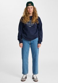 Tengerészkék pulóver fehér logóval, laza szabású világoskék farmernadrág, és szürke-barna sneakers, zöld sapkával kiegészítve.