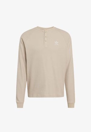 Beige pitkähihaine henley-paita, jossa on teksturoitu kangas, kolmen napin lista ja valkoinen logo vasemmassa rinnassa. Ribatut hihansuut ja kaulus.