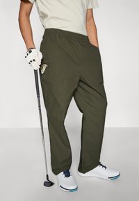 Pantalon de golf vert olive avec une ceinture élastique, des poches latérales et un logo brodé sur la cuisse gauche. Associé à des chaussures de golf blanches.