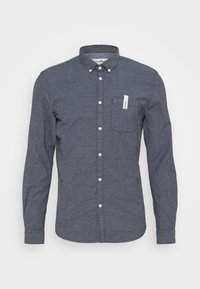 Camisa de botón azul oscuro hecha de una tela texturada, con un bolsillo en el pecho y botones blancos en la parte delantera. Diseño de cuello clásico.