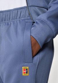 Nike träningsoveraller i ljusblått. Har texturerat tyg, elastisk midja, sidoficka och en flerfärgad broderad logopatch.
