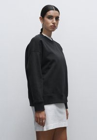 Sudadera negra con cuello redondo y bajo acanalado, combinada con una mini falda blanca. Presenta mangas largas y un ajuste relajado. Diseño sencillo.