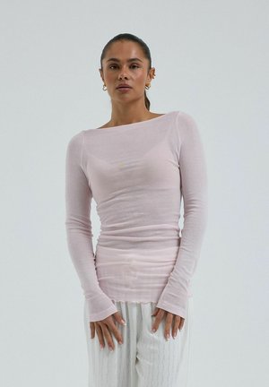 Femme aux cheveux attachés, portant un haut transparent à manches longues rose pâle et un pantalon blanc à motifs, debout devant un fond uni.