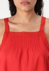 Top roșu fără mâneci, cu un guler dantelat, design cu pliuri în față și material ușor. Textura pare moale și netedă.