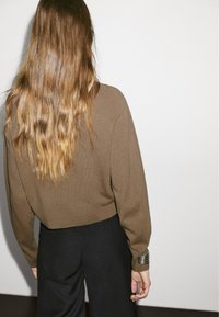 Brown knit sweater with long sleeves and a cropped fit, paired with wide black trousers. Notable silver bracelet on wrist.