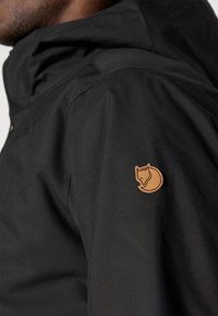 Melna ūdensnecaurlaidīga jaka ar teksturētu virsmu, kurai uz kreisā pleca ir brūna lapsas logotipa uzlīme. Ar kapuci un pogu detaļām.