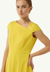 Gelbes Kleid mit Cap-Ärmeln, das ein diagonales Nahtdetail und eine glatte Textur aufweist. Das Model trägt goldfarbene Ohrringe.