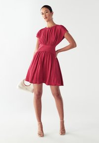 Czerwona sukienka o krótkim rękawie z dopasowaną talią i rozkloszowanym dołem. Materiał wydaje się lekki i fakturowany, w połączeniu z przezroczystymi sandałami na obcasie i białą torebką.