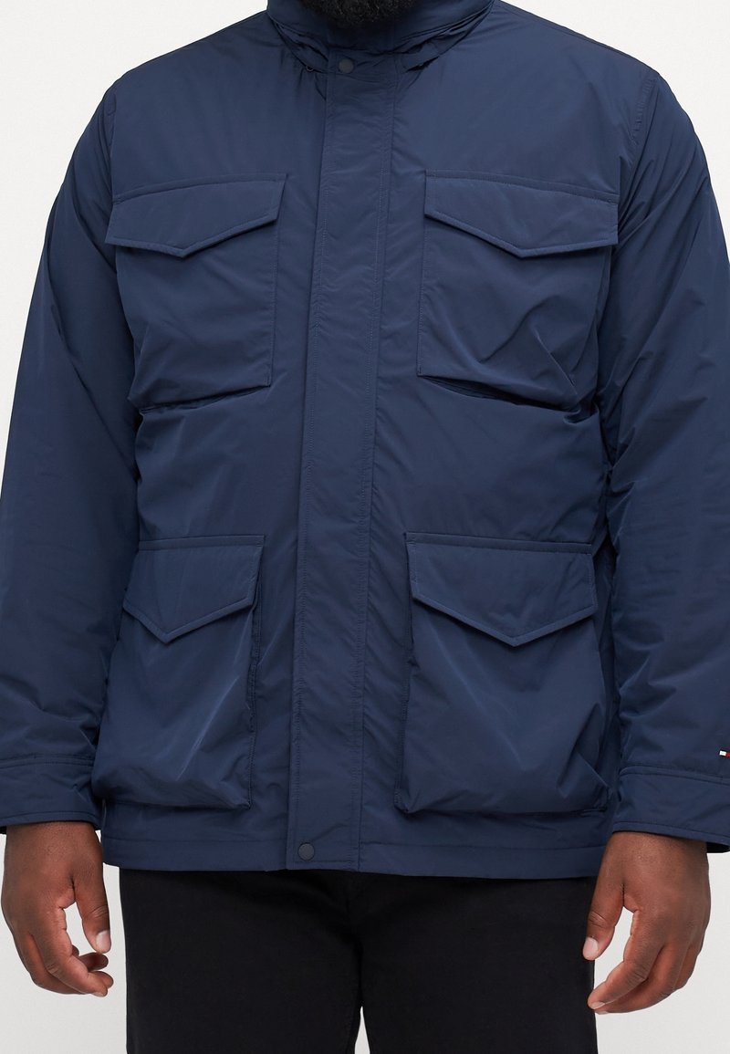 Homme portant une veste bleu marine avec quatre poches à rabat à l'avant et un pantalon noir, debout les bras détendus le long du corps.