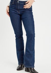 Dunkelblaue Jeans mit Schlag, ausgestattet mit kontrastierendem Nähen, im Fünf-Taschen-Stil und mit Reißverschluss, kombiniert mit schwarzen Knöchelstiefeln.