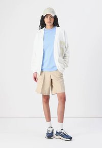Cardigan blanc avec des accents beiges, T-shirt bleu clair, shorts beiges et baskets navy. Le mannequin porte une casquette claire et des chaussettes blanches.