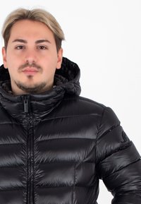 Černá péřová bunda s lesklým povrchem, horizontální prošívaný vzor, zipový uzávěr a nastavitelná kapuce. Na rukávu má logo.