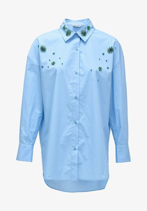 Camisa abotonada de color azul claro con adornos florales en verde esmeralda y pequeños remaches. Presenta un collar clásico y mangas largas.