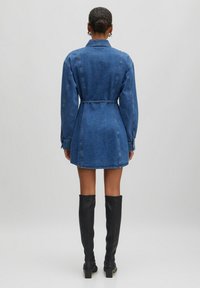 Denimhemdjurk in donkerblauw, met lange mouwen en een ceintuur in de taille. Gladde textuur met een rechte snit, gecombineerd met kniehoge zwarte laarzen.