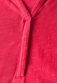Tissu rouge avec une finition douce et texturée. Présente un col en V avec des coutures visibles, créant un design épuré et simple.