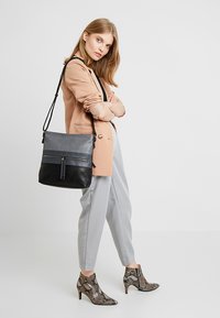 Bolso de hombro de cuero gris y negro con un bolsillo frontal, cierre de cremallera y un acento de borlas. Correa ajustable; diseño elegante y moderno.