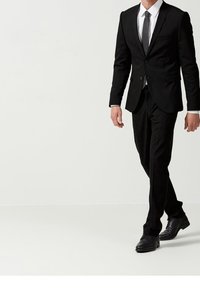 Traje negro con solapas, camisa de vestir blanca y corbata negra estrecha. La persona lleva zapatos de vestir negros pulidos, caminando sobre una superficie clara.