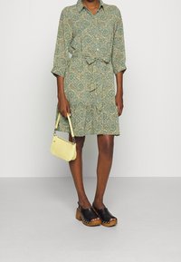 Viherääkuvioinen paidan mekko, jossa on solmittava vyötärö ja kolmanneksen pituiset hihat. Yhdistetty mustiin puukengiin ja pieneen keltaiseen olkalaukkuun.