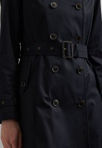 Marine trenchcoat met dubbele knopen, een ceintuur in de taille en discrete zakken. Het gladde materiaal en de accenten in goudkleur verbeteren het ontwerp.