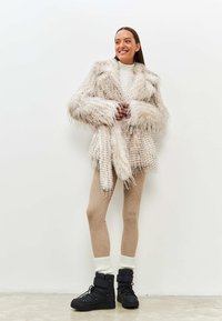 Lang faux fur frakke i lys beige med shaggy tekstur, parret med ribbede beige leggins og sorte snørebåndsstøvler med tykke såler.