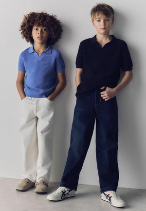 Kaksi poikaa seisoo seinää vasten, toinen sinisessä lyhythihaisessa neuleessa valkoisissa housuissa, toinen mustassa lyhythihaisessa neuleessa tummissa farkuissa.