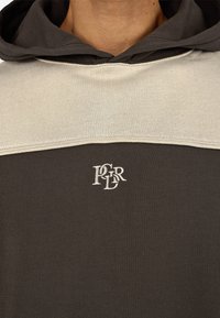 Hættebluse i mørkebrun og beige. Har kontrasterende farveblokke, blødt stof og et lille hvidt logo på brystet.