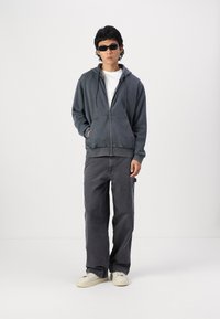 Gray zip-up hoodie with a front pocket, white t-shirt underneath, dark gray pants, and white sneakers; model wearing black sunglasses.