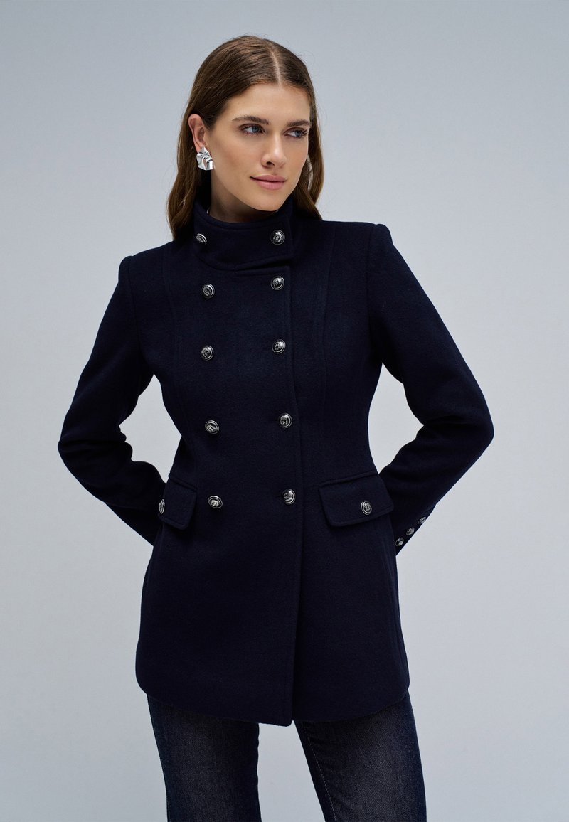 Donna che indossa un cappotto doppiopetto aderente blu navy con bottoni d'argento, in piedi con le mani dietro la schiena contro uno sfondo chiaro semplice.