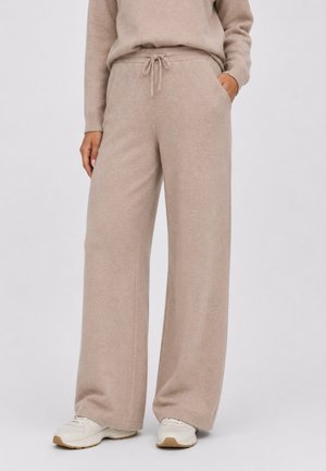 Person iført lyse beige vidde strikkede bukser med snøre i taljen og cremehvide sneakers står mod en ensfarvet baggrund.