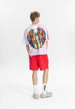 Joven de espaldas con una camisa tie-dye en tonos pastel con alas de lentejuelas de colores, pantalones cortos rojos, calcetines blancos y zapatos beige texturizados.