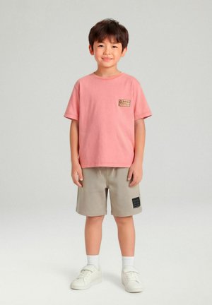 Garçon debout et souriant portant un t-shirt rose, un short beige, des chaussettes blanches et des baskets blanches sur un fond gris uni.