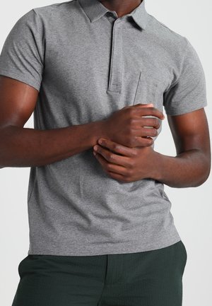 Man wearing a short-sleeve gray polo shirt and dark green pants, with hands clasped at waist against a plain background.