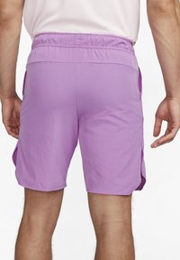 Man klädd i lila atletiska shorts med elastiskt midjeband och sidofickor, som står framför en vit bakgrund.