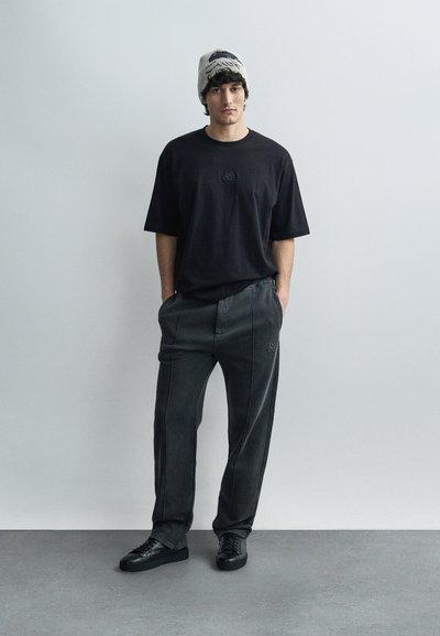 Black oversized t-shirt with a small logo, paired with dark, tailored pants featuring contrast stitching, and black sneakers. Grey beanie included.