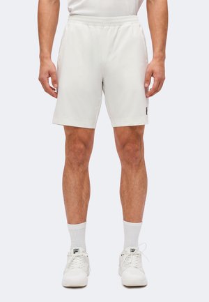 Mand iført hvide atletikshorts, hvide sokker og hvide sneakers, stående med benene let spredt mod en ensfarvet baggrund.