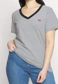 Žena v černobílem pruhovaném tričku s výstřihem do V od Levi's a modrých džínách, ruka zastrčená v kapse na jednobarevném pozadí.