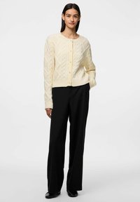Cardigan a maglia cable crema con scollo rotondo e chiusura con bottoni, caratterizzato da motivi testurizzati. Abbinato a pantaloni neri a gamba larga.