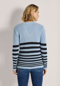 Frau mit schulterlangem blonden Haar, die einen hellblauen Strickpullover mit schwarzen horizontalen Streifen trägt, steht vor einem schlichten hellen Hintergrund.